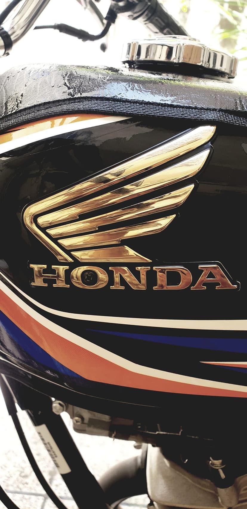 Golden Honda wing logo on a black motorcycle tank with orange and blue racing stripes and chrome fuel cap.