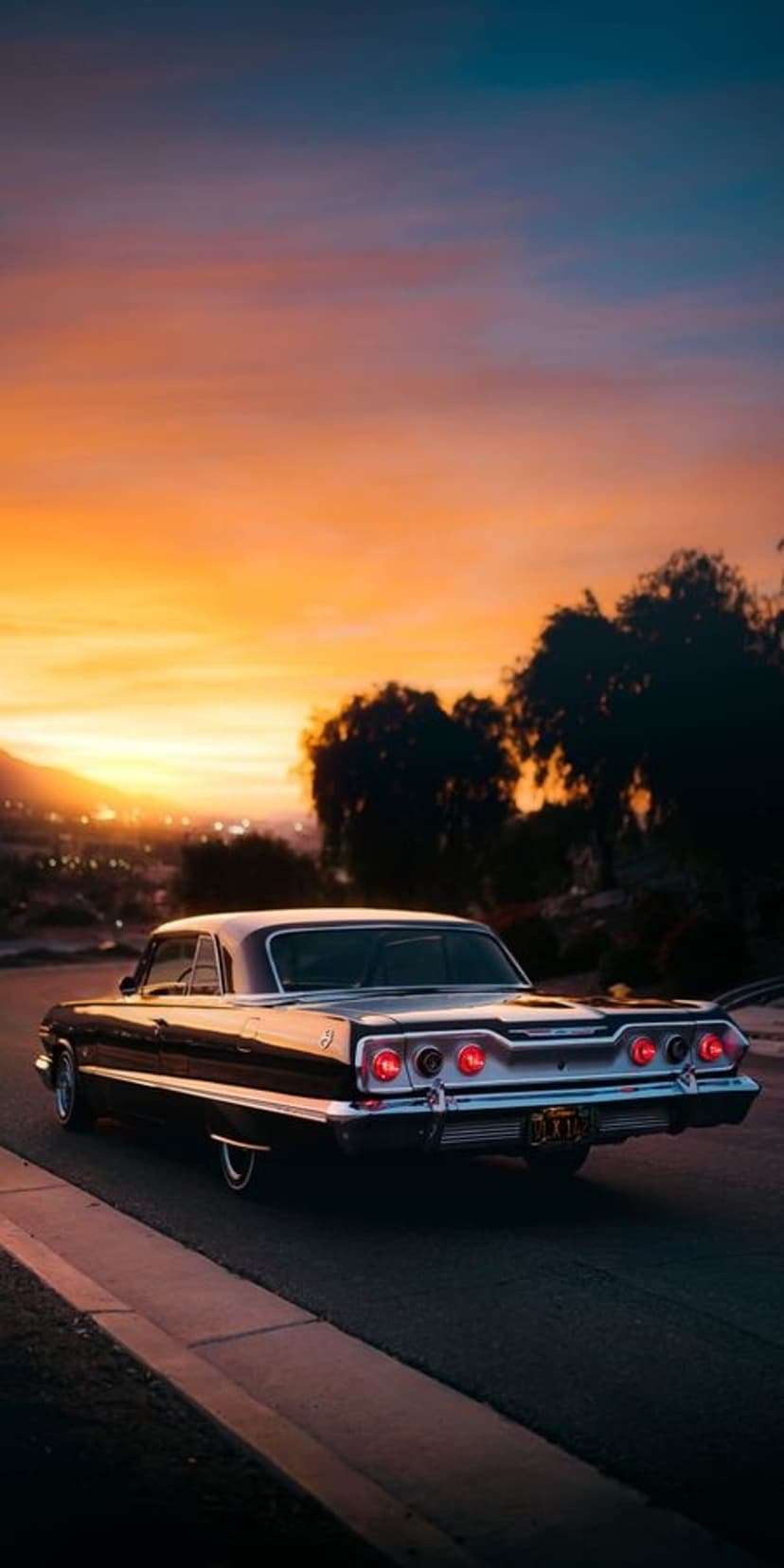 Black Chevrolet Impala with glowing red brake lights drives on a road under a vibrant orange and blue sunset.