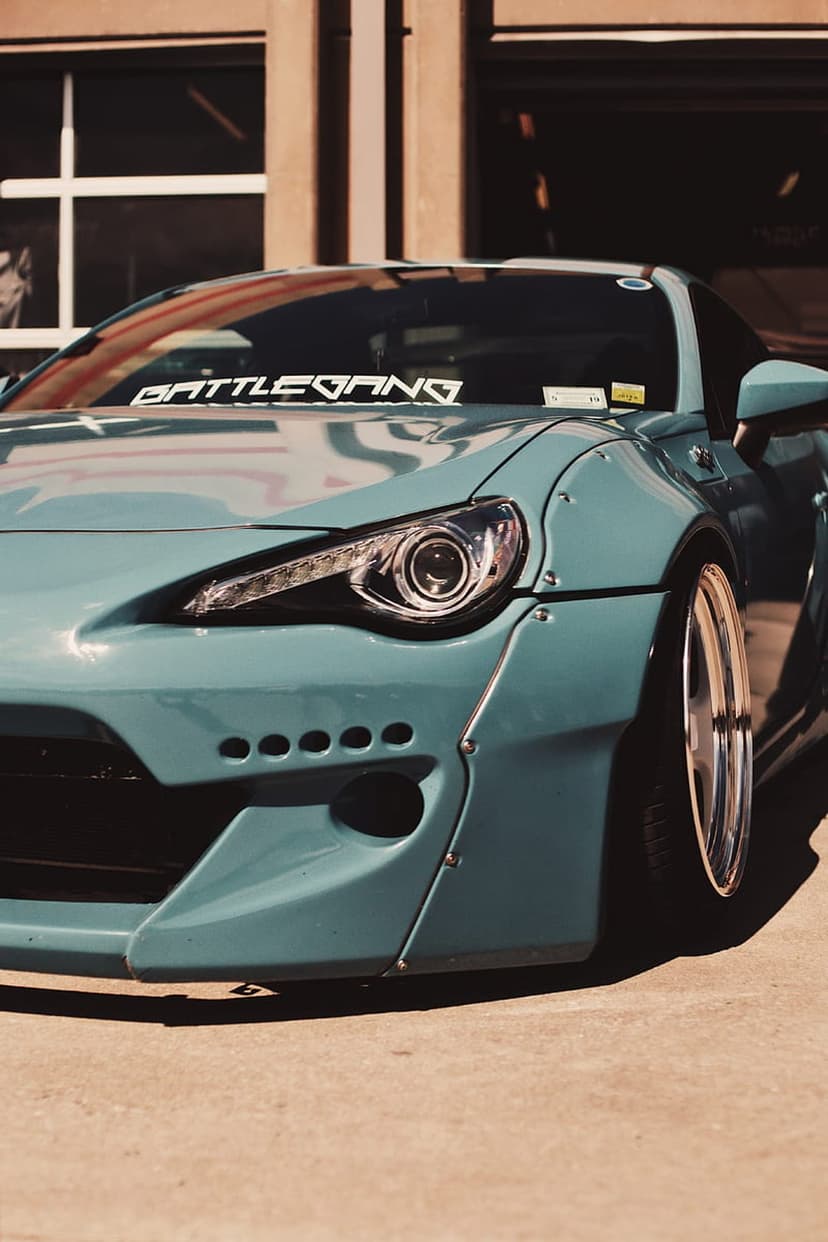 Teal widebody car with a Battle Gang windshield banner, flared fenders, chrome wheels, and aggressive front bumper.