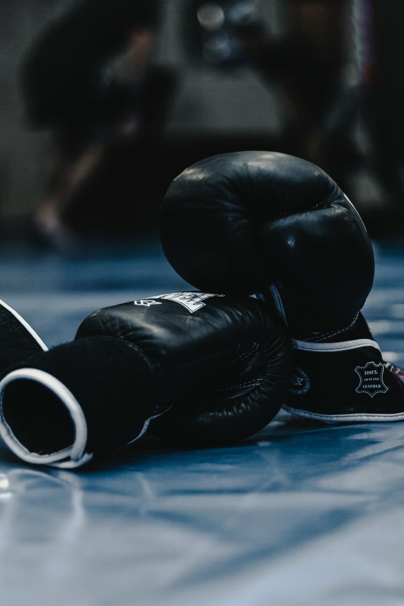 Black boxing gloves with white stitching rest on a textured blue mat inside a gym with a blurred background.