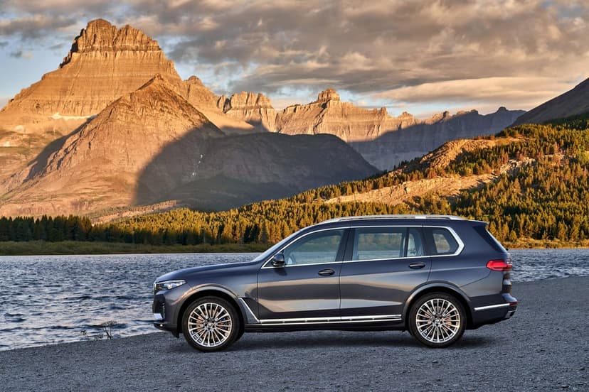 Dark gray luxury SUV parked on a gravel lake shore with towering golden mountains in the background.