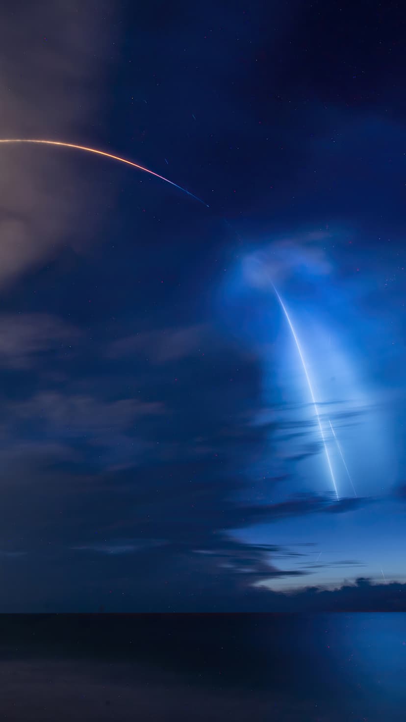 Rocket launch plume glowing blue above a dark ocean horizon with a bright white light trail in the night sky.