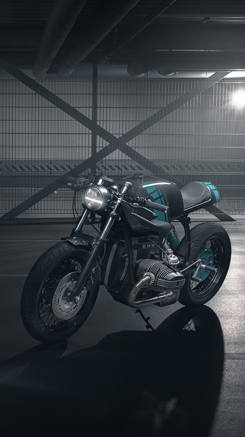 Black cafe racer motorcycle with teal accents parked on a reflective concrete floor in a dark industrial garage.