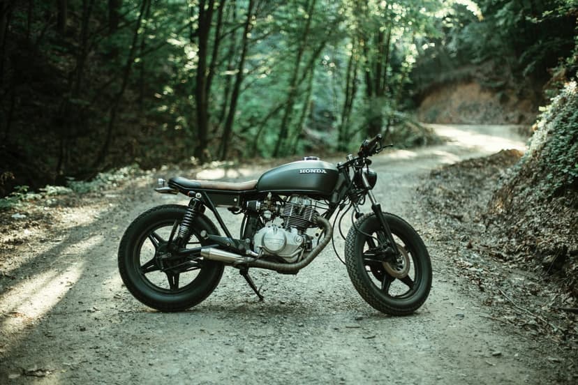 Black cafe racer motorcycle parked on a dirt trail beneath a sunlit forest canopy with vibrant green trees.