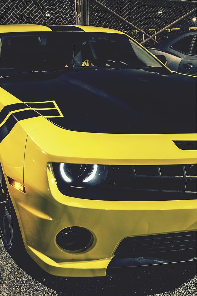 Yellow Chevrolet Camaro with black racing stripes and glowing circular LED halo headlights in a front view.