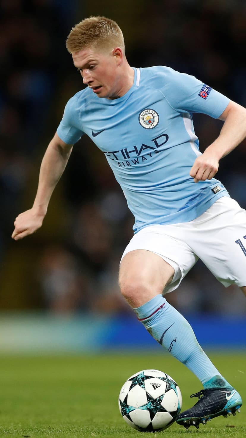 Kevin De Bruyne in a light blue Manchester City kit dribbling a football on a green grass pitch during a match.