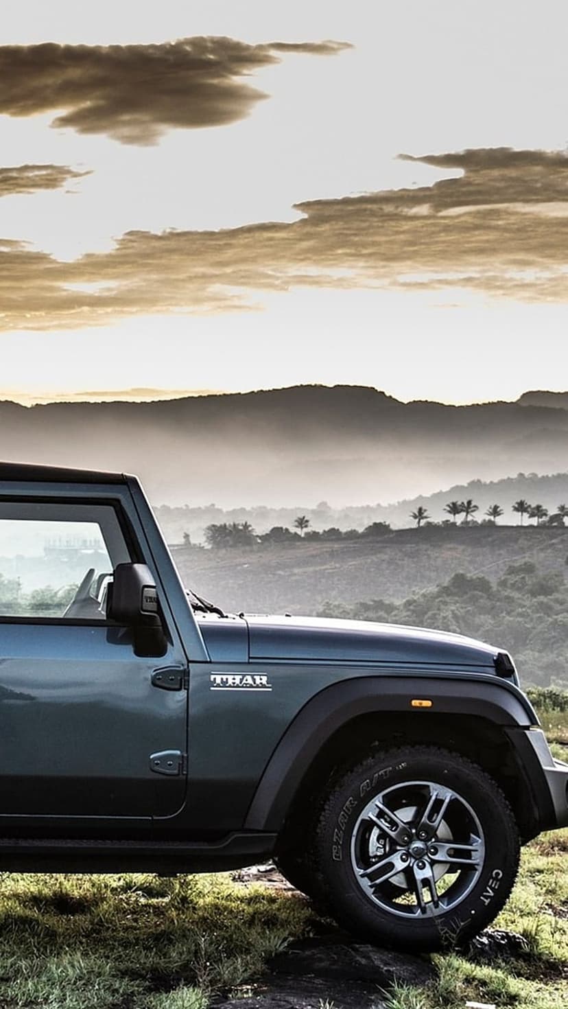 Dark blue Mahindra Thar SUV parked on a grassy hill with hazy mountains and a sunset sky in the background.