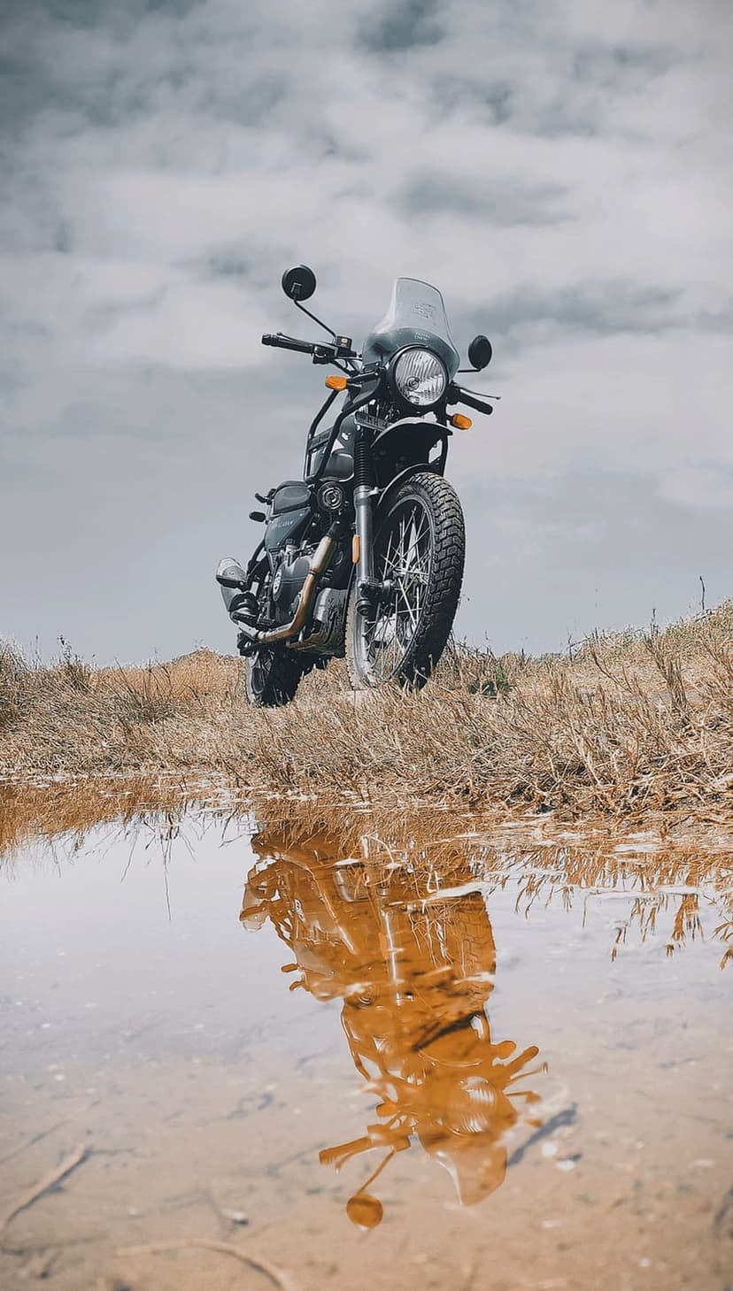 Adventure motorcycle with knobby tires parked on dry grass reflects in a large puddle under a gray sky.