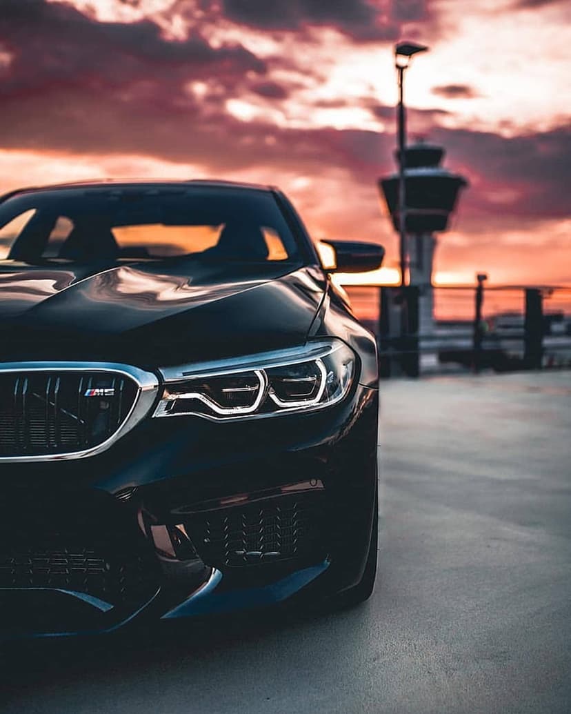 Black BMW M5 sport sedan with lit headlights parked at twilight near an airport control tower silhouetted by clouds.
