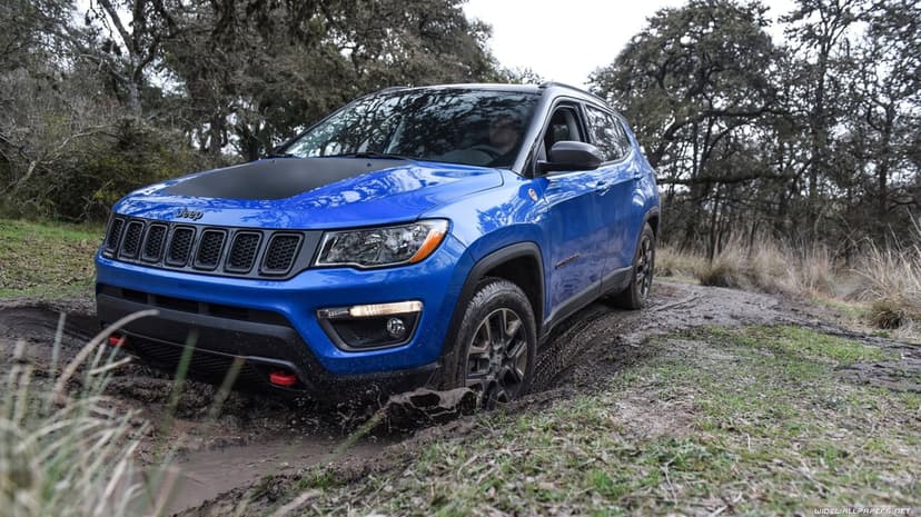 Blue Jeep Compass Trailhawk splashing through deep mud on a wooded trail with a matte black hood and red tow hooks.