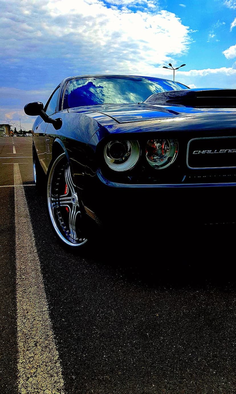 Dark blue Dodge Challenger R/T parked on asphalt with chrome wheels reflecting a bright cloudy sky at low angle.