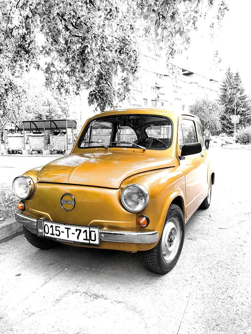 Yellow vintage Fiat 600 car parked on a city street next to silver metal trash bins under a leafy green tree.