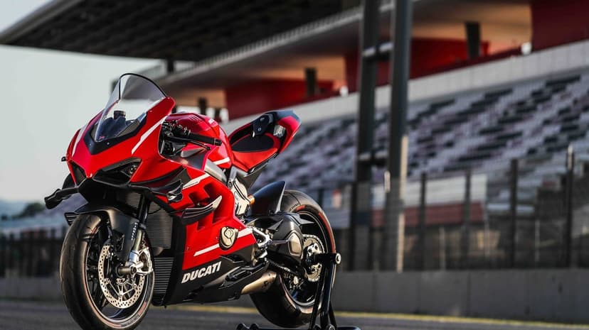 Red Ducati sportbike with carbon fiber wings on a paddock stand at a sunlit racetrack with stadium seating.