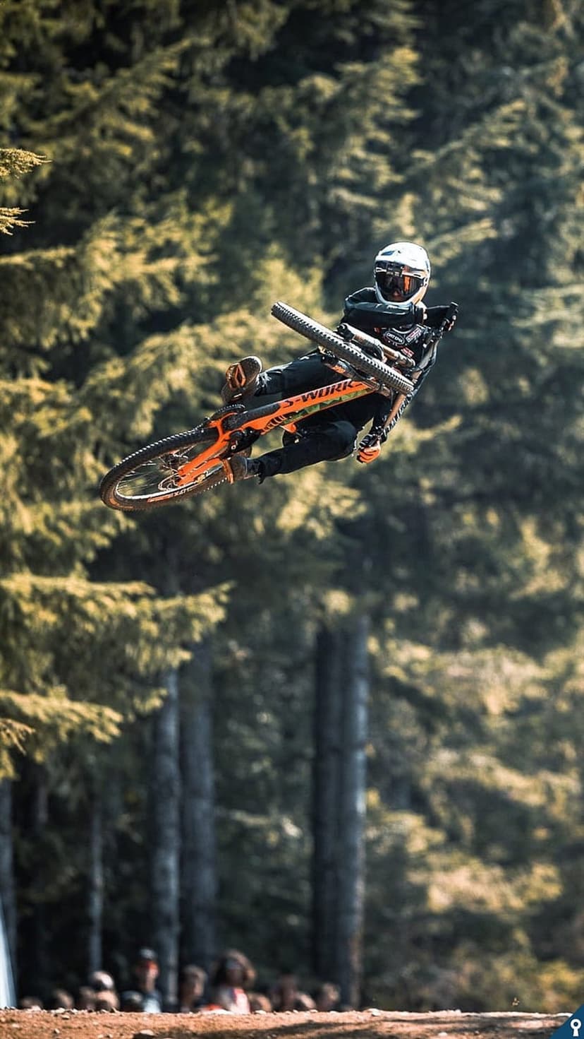 Mountain biker soaring mid-air over a dense evergreen forest during an extreme downhill bike competition.