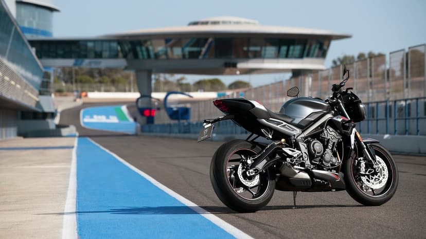 Silver Triumph motorcycle on a racetrack with blue rumble strips and a futuristic tower under a clear sky.