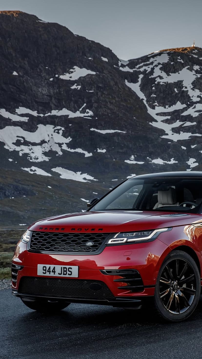 Red Range Rover Velar parked on a snowy mountain road with rugged alpine peaks under a cloudy winter sky.