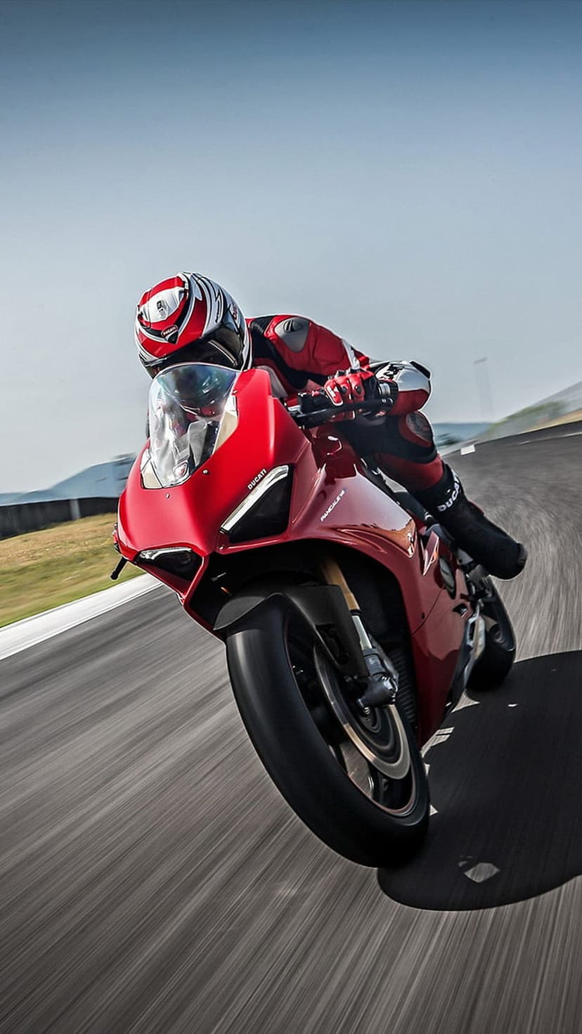 Red Ducati rider leaning into a sharp racetrack curve with blurred asphalt and a clear blue sky background.