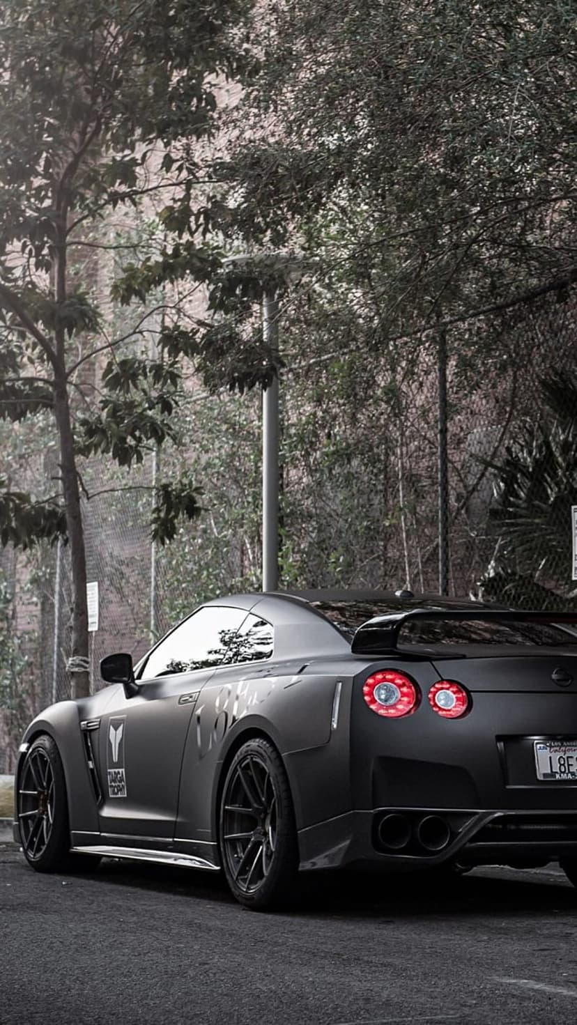 Nissan GT-R in matte black features red taillights and custom wheels on a city street with green trees behind it.