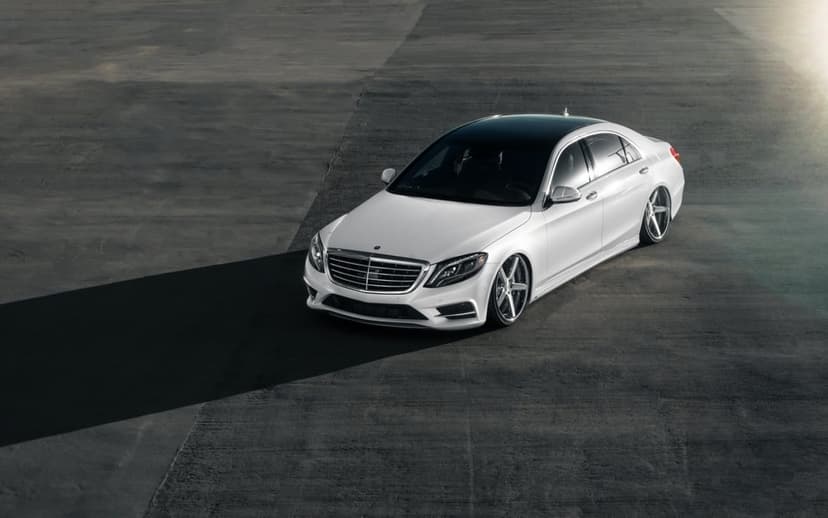White Mercedes-Benz S-Class sedan with chrome wheels and black roof on gray concrete with a long shadow.