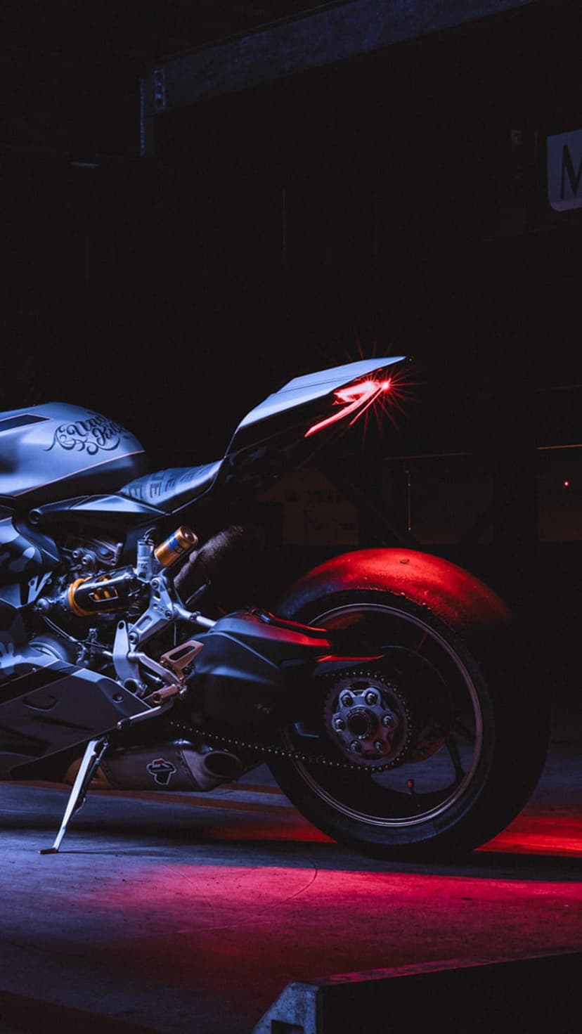 Ducati motorcycle rear tail light glows red in a dark garage, illuminating the tire and suspension components.