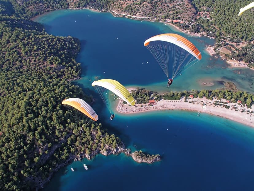 Three paragliders with red and blue canopies fly over a turquoise bay and a green coastline with a sandy beach.