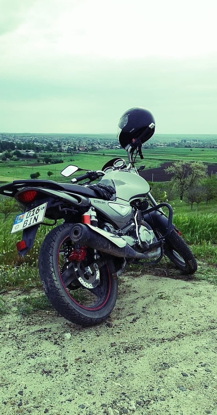 Motorcycle with a black helmet on the handlebars parked on a dirt path overlooking a green valley and town.