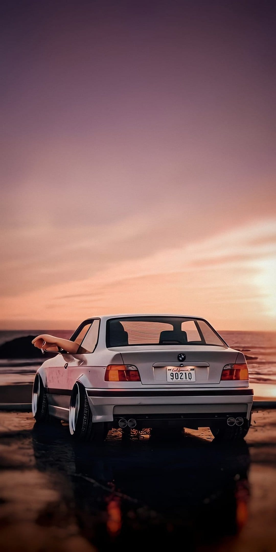 BMW E36 convertible rear profile parked on a wet beach with taillight reflections during a vibrant sunset.