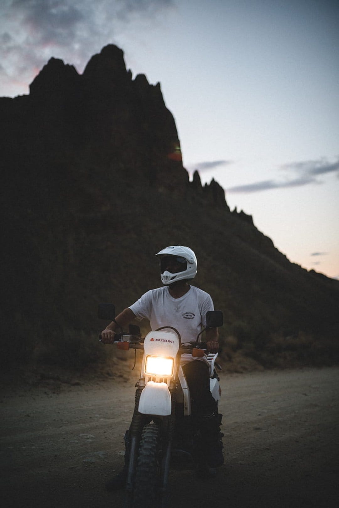 Motorcyclist in a white helmet sits on a white dirt bike with its headlight on before dark mountain silhouettes.