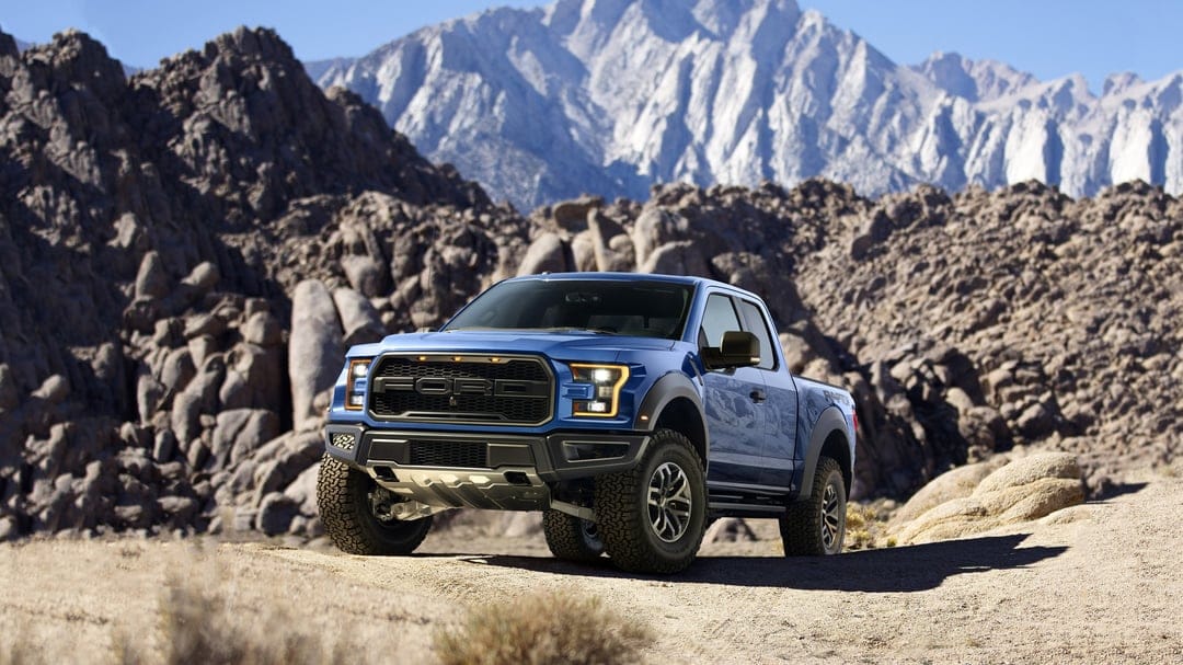 Blue Ford Raptor pickup truck parked on a rocky desert trail with jagged mountain peaks in the background.