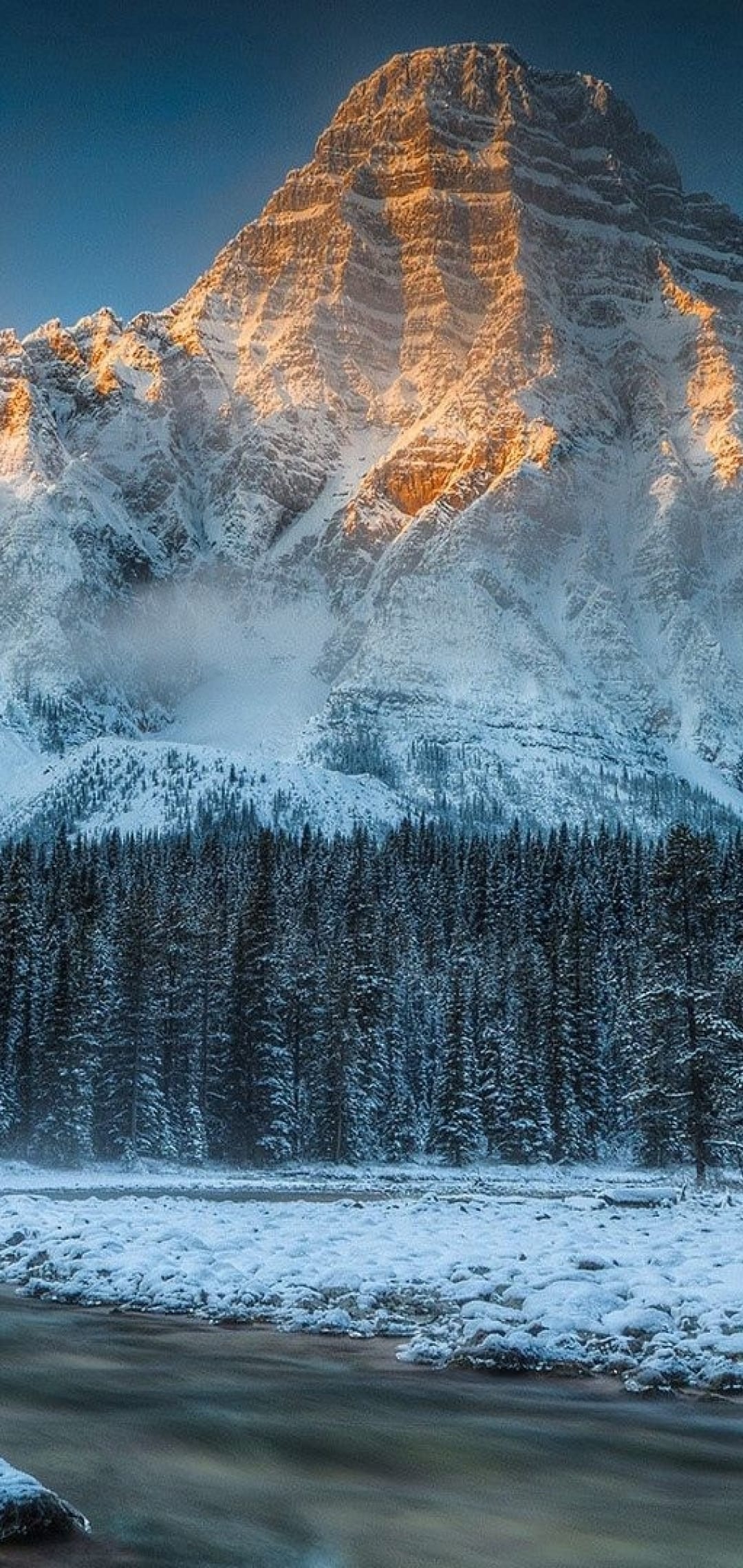 Snow-capped mountain peak glowing with golden sunlight above a frozen river and evergreen forest in winter.