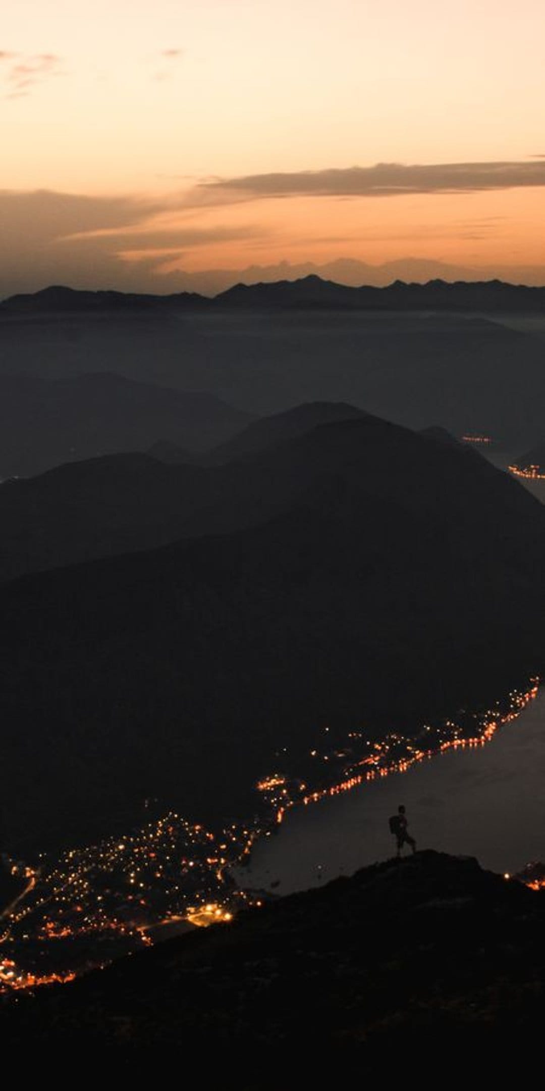 Solitary person stands on a high cliff overlooking a glowing lakeside town nestled between dark mountain peaks.