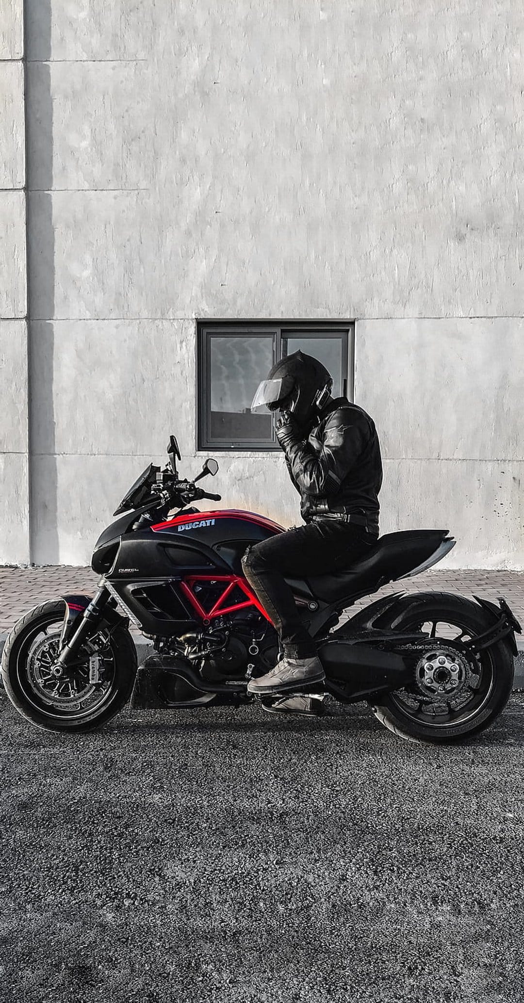 Rider wearing a black leather jacket and helmet sits on a red and black Ducati motorcycle against a white wall.