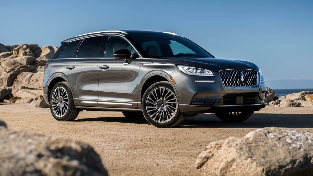Charcoal gray luxury SUV parked on coastal sands with light rocks and a clear blue sky background.