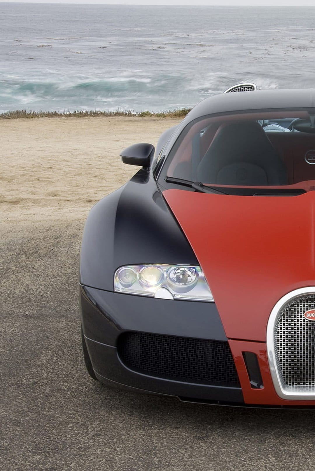 Bugatti Veyron in red and black parked on a shoreline with ocean waves crashing in the blurred background.