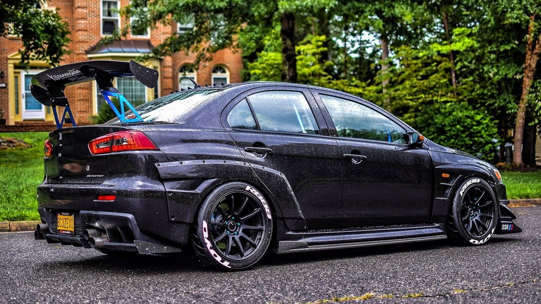 Modified black Mitsubishi Lancer Evolution X with a widebody kit and large rear wing parked on a wet street.