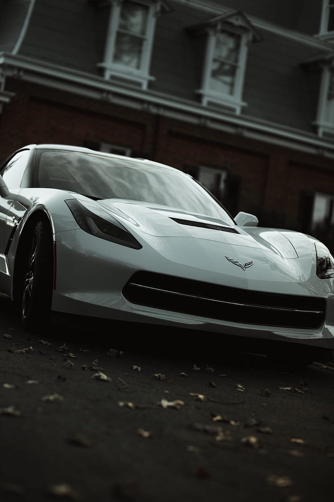 White Chevrolet Corvette C7 parked on dark asphalt with autumn leaves near a rustic red brick building facade.