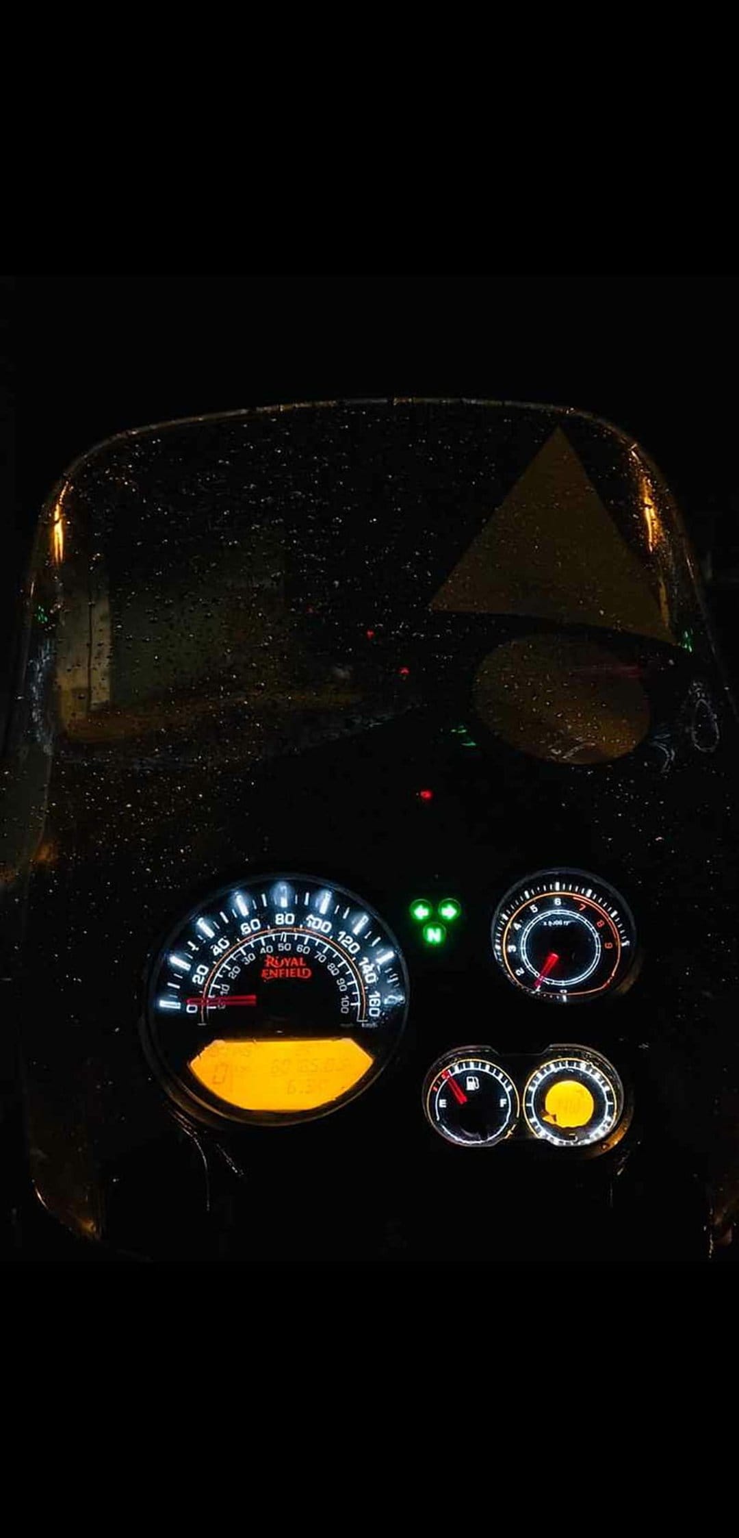 Royal Enfield Himalayan motorcycle dashboard glowing with amber light behind a rain-streaked windscreen at night.