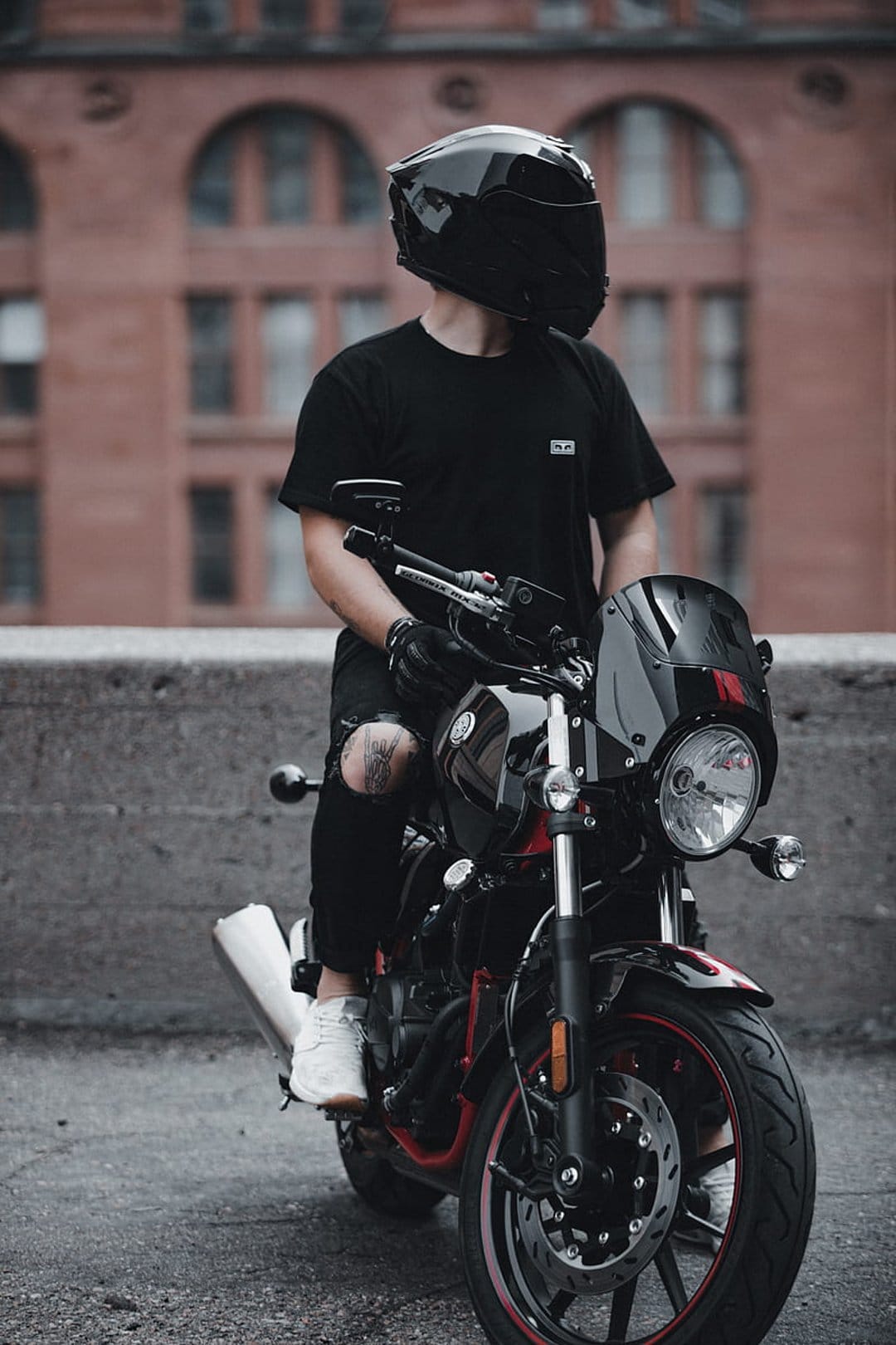 Biker in a black helmet and ripped jeans sits on a motorcycle with red accents against a brick wall.