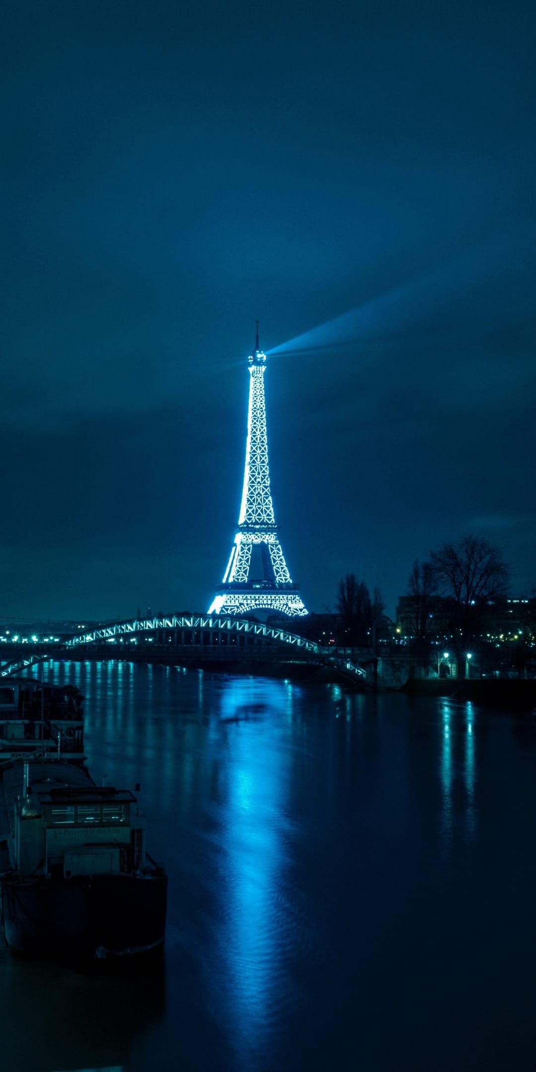 Eiffel Tower illuminated with bright blue neon lights against a dark night sky with river reflections below.