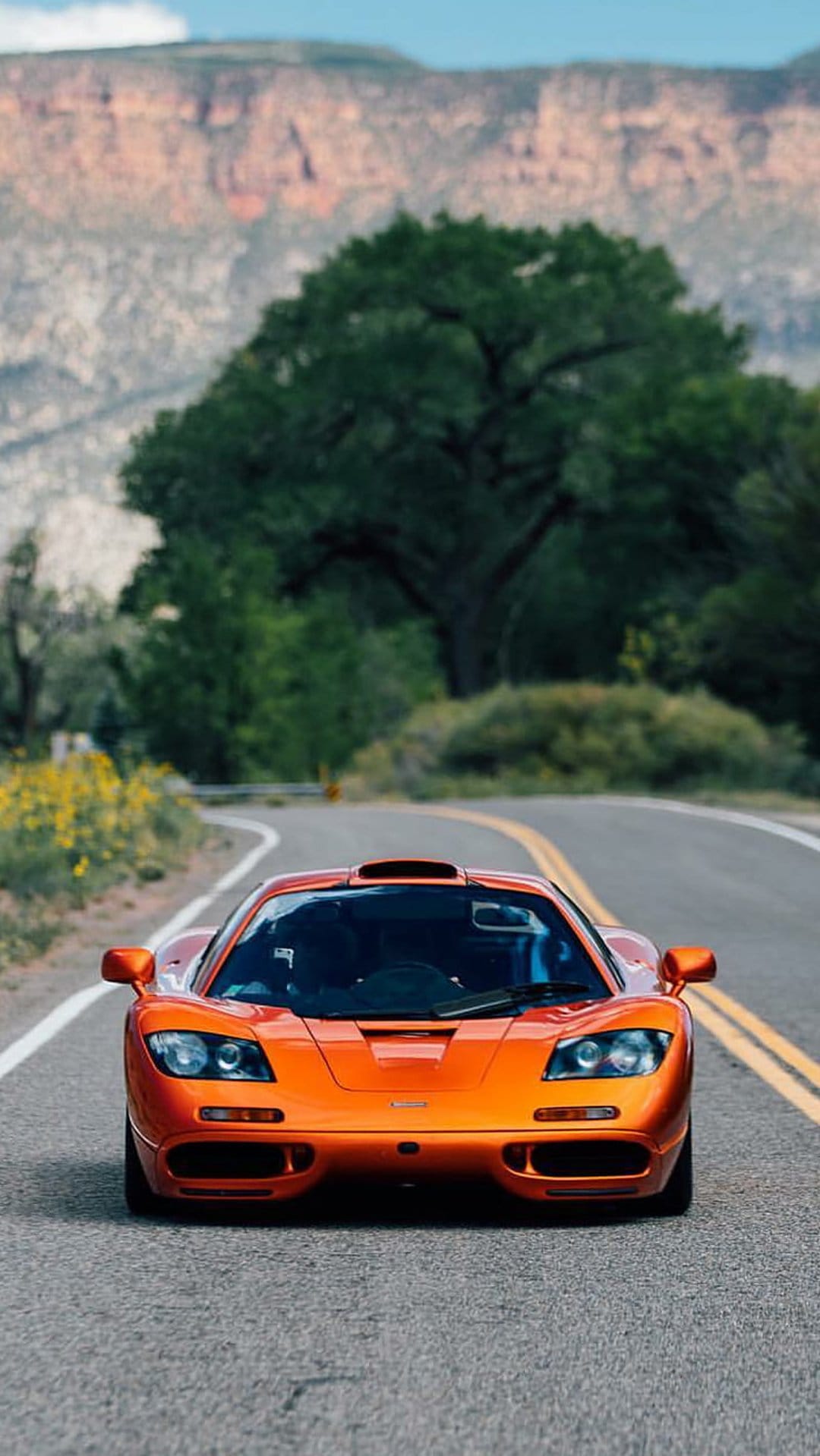 Orange McLaren F1 supercar driving on a winding desert road with mountains and green trees in the background.
