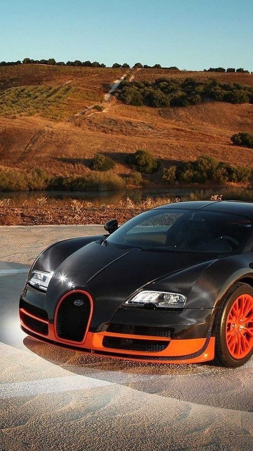 Black Bugatti Veyron with orange wheels and grille drives on sand near a lake under a bright blue summer sky.