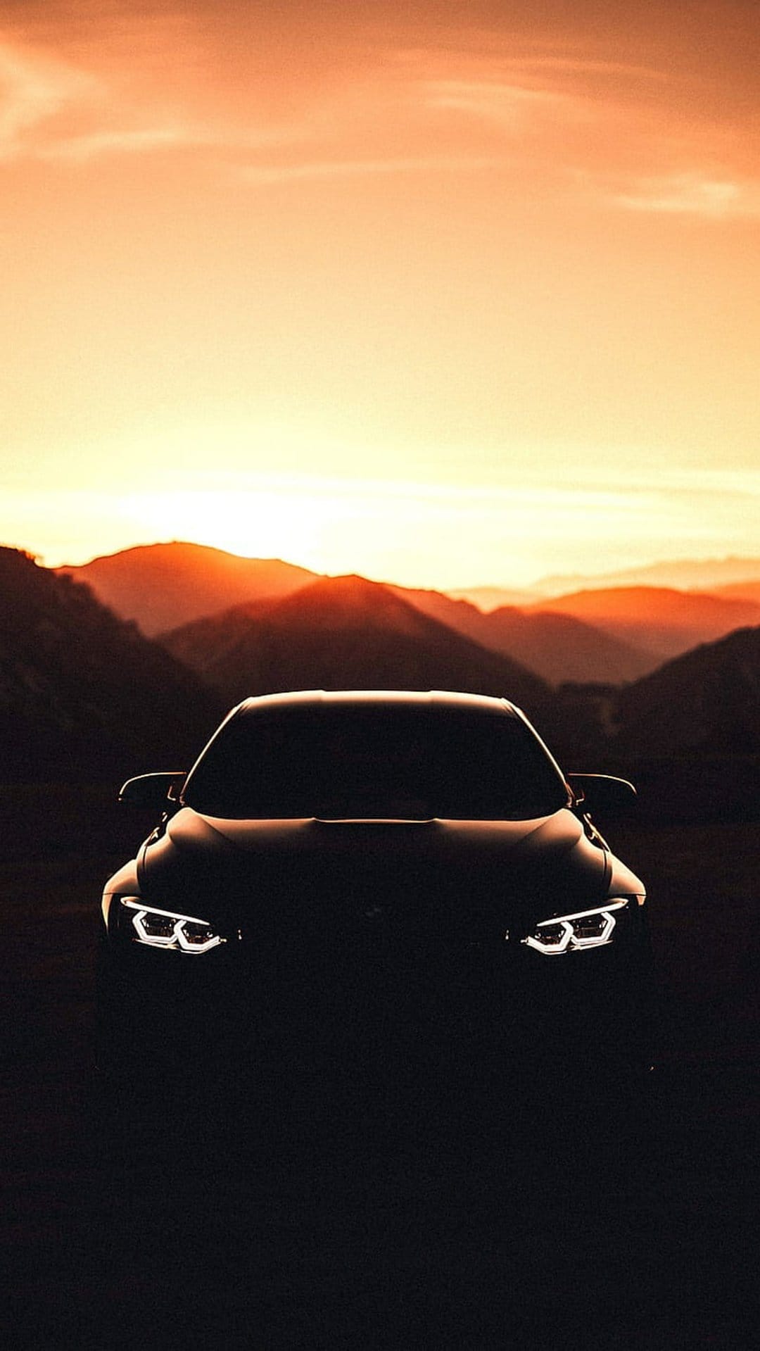 Glowing car headlights shine on a dark silhouette with layered mountains under an orange sunset sky.