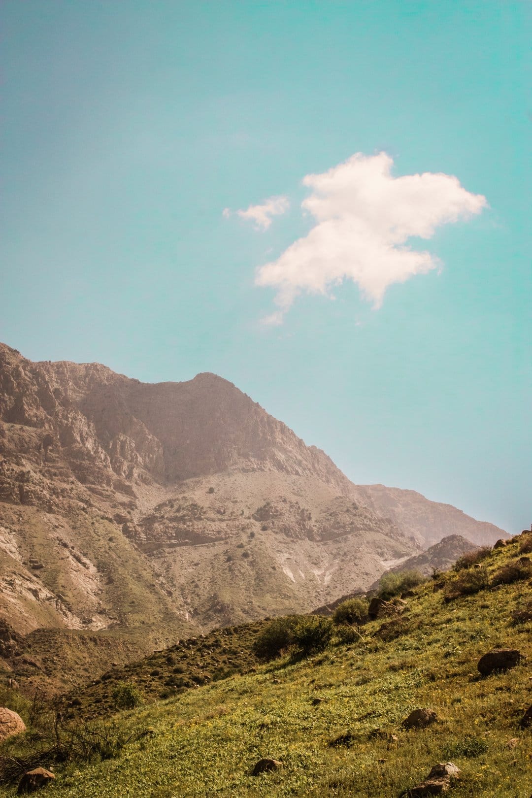 Rugged mountain peak against a turquoise sky with a single cloud and a green grassy slope in the foreground.