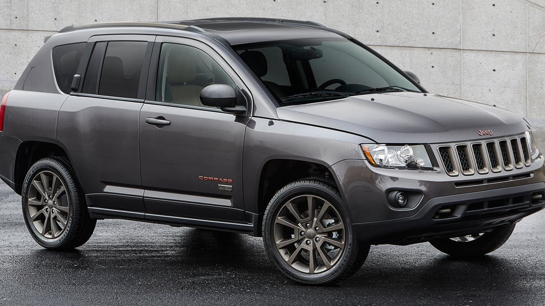 Dark grey Jeep Compass SUV with bronze wheels parked on wet asphalt in front of a textured concrete wall.