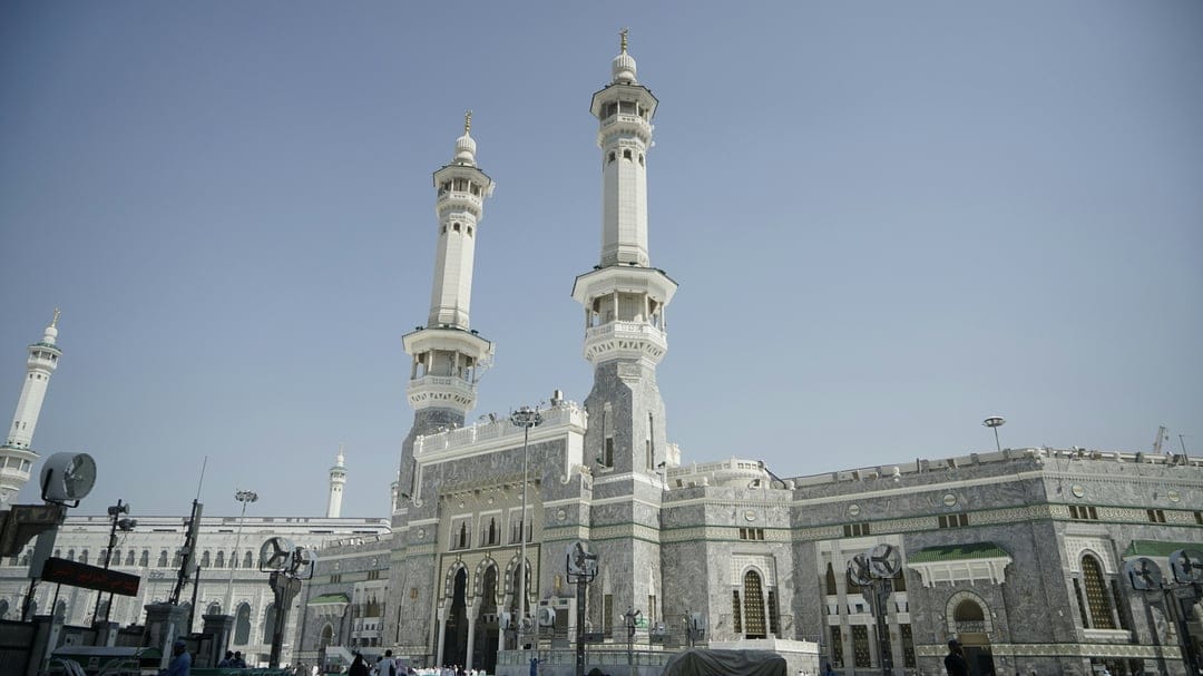 Two stone minarets of Masjid al-Haram tower over a low-angle view of the mosque against a clear blue sky.
