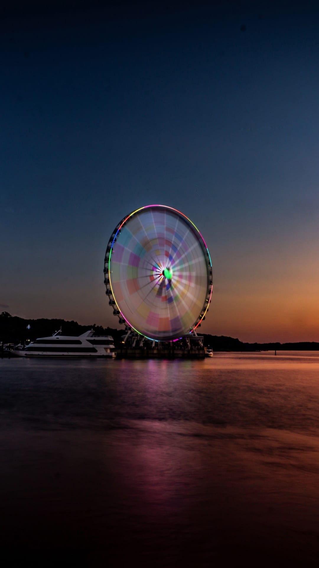 Illuminated Ferris wheel with colorful light streaks reflects on dark water beneath a gradient twilight sky.