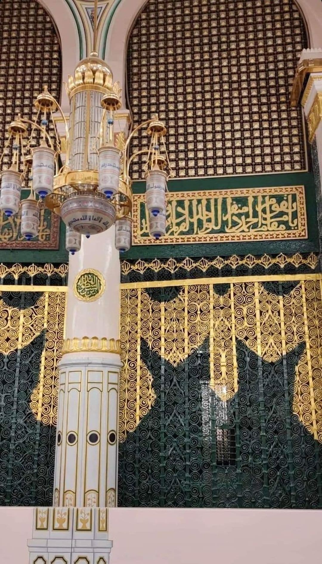 Golden mosque chandelier with cylindrical lamps and Arabic calligraphy against green latticework screens.