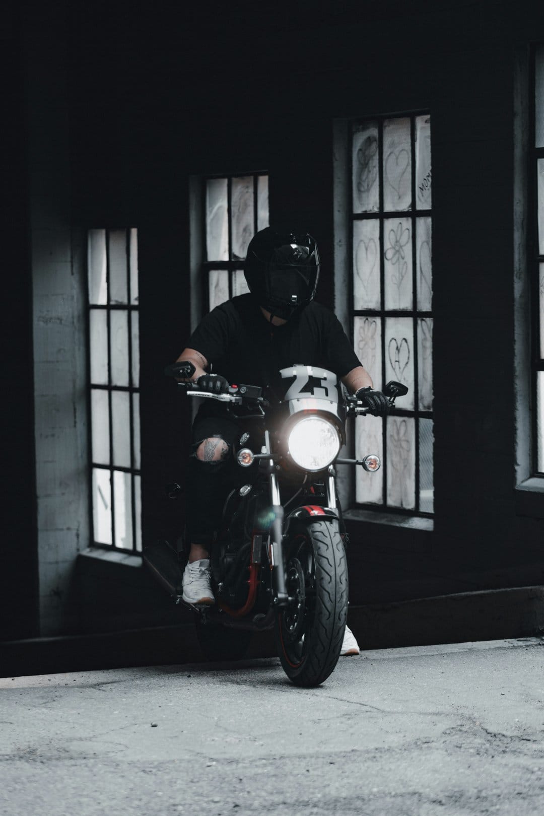 Motorcyclist in a black helmet and ripped jeans rides through a dark graffiti alley with a glowing headlight.