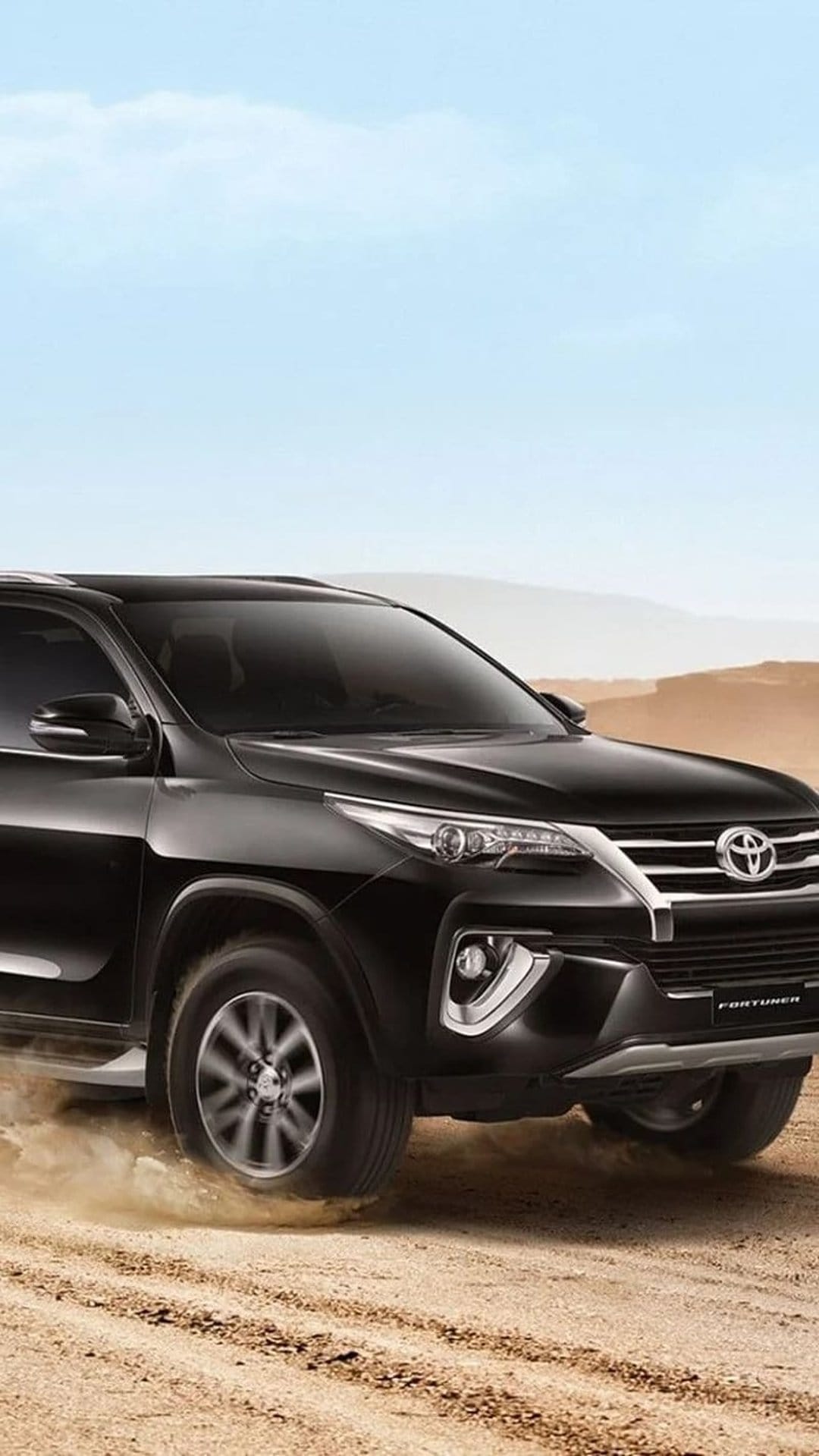 Black Toyota Fortuner SUV driving through dusty desert dunes under a clear blue sky on a vertical screen.