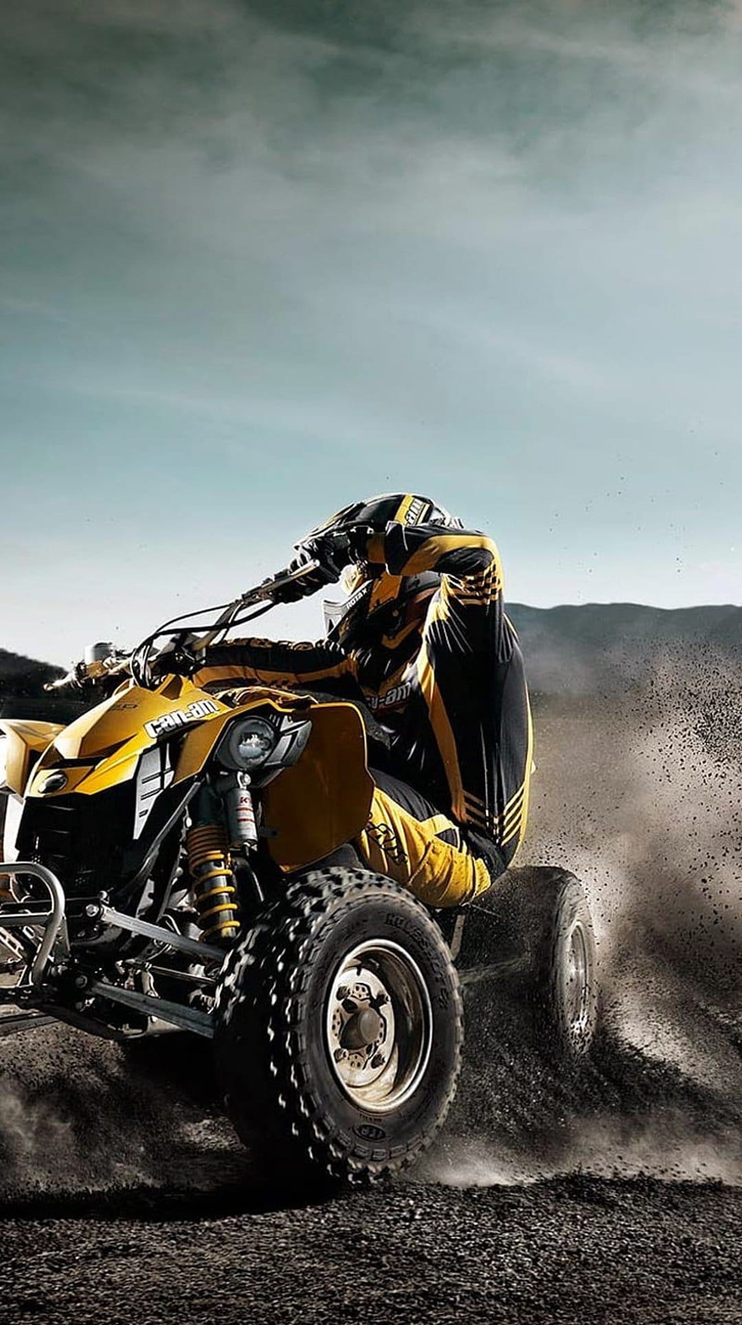 Rider in yellow gear leans a yellow quad bike into a sharp dirt turn, kicking up dust under an overcast sky.